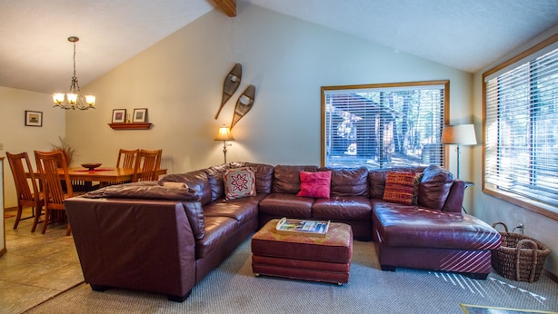 A living room with a sectional sofa, ottoman, dining table, chairs, chandelier, wall decor, and large windows with blinds.