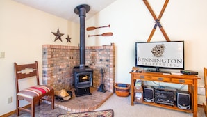 A cozy living room with a brick fireplace, a wooden chair, a television, and a wooden table with speakers.