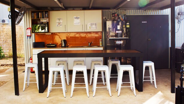 A kitchen with a long counter, white stools, and a refrigerator. - Down Under Hostels on Harriet (Darwin)
