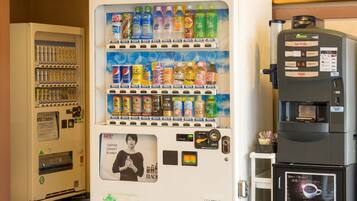 Máquina expendedora con varias bebidas y aperitivos, una cafetera y un refrigerador con publicidad de café.