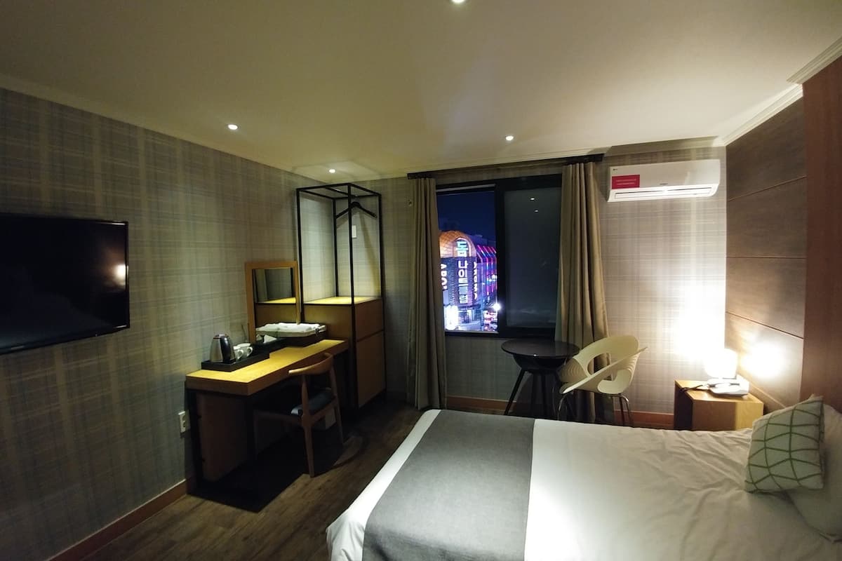 A hotel room with a bed, a desk with a chair, a television, and a window with a view of a building at night.