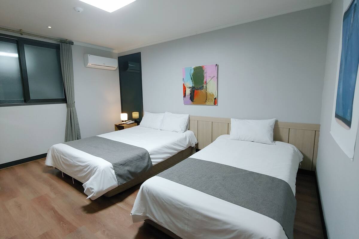 A hotel room with two beds, a window with curtains, an air conditioning unit, a painting on the wall, and a bedside table with a lamp.