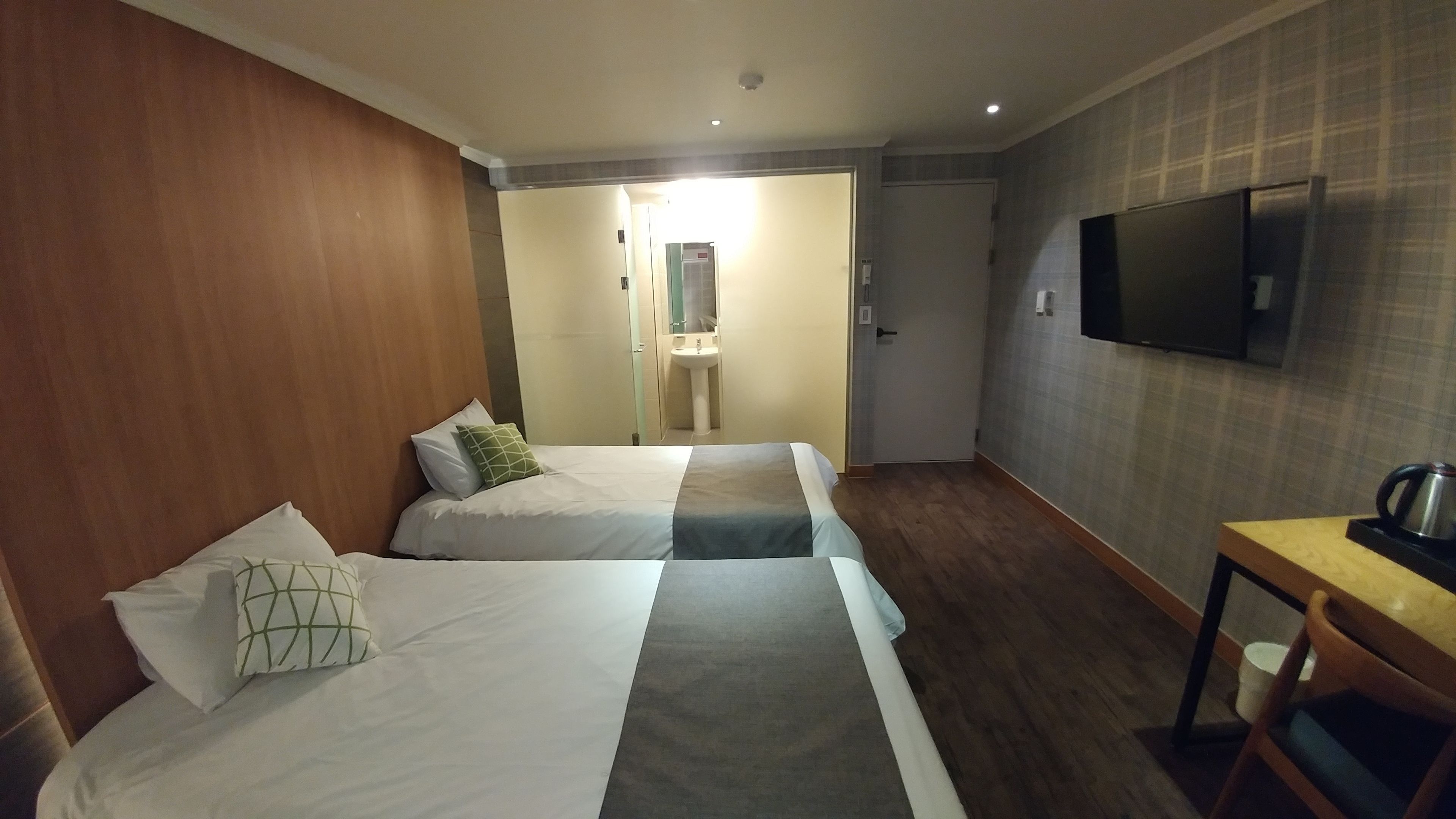 A hotel room with two beds, a TV, a table with a kettle, and a bathroom visible through an open door.