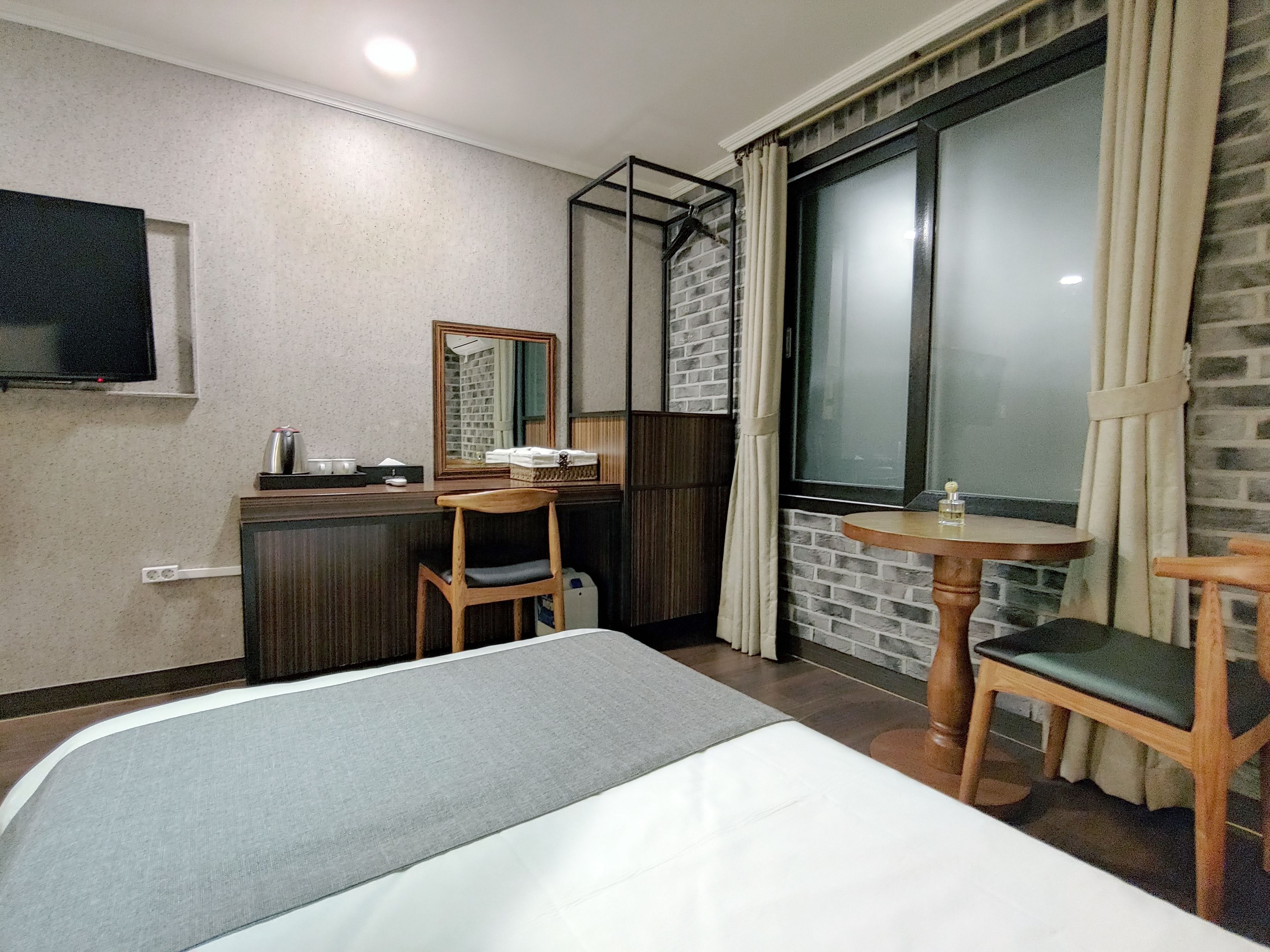 A hotel room with a bed, a desk with a chair, a small table, a TV mounted on the wall, and a window with curtains.