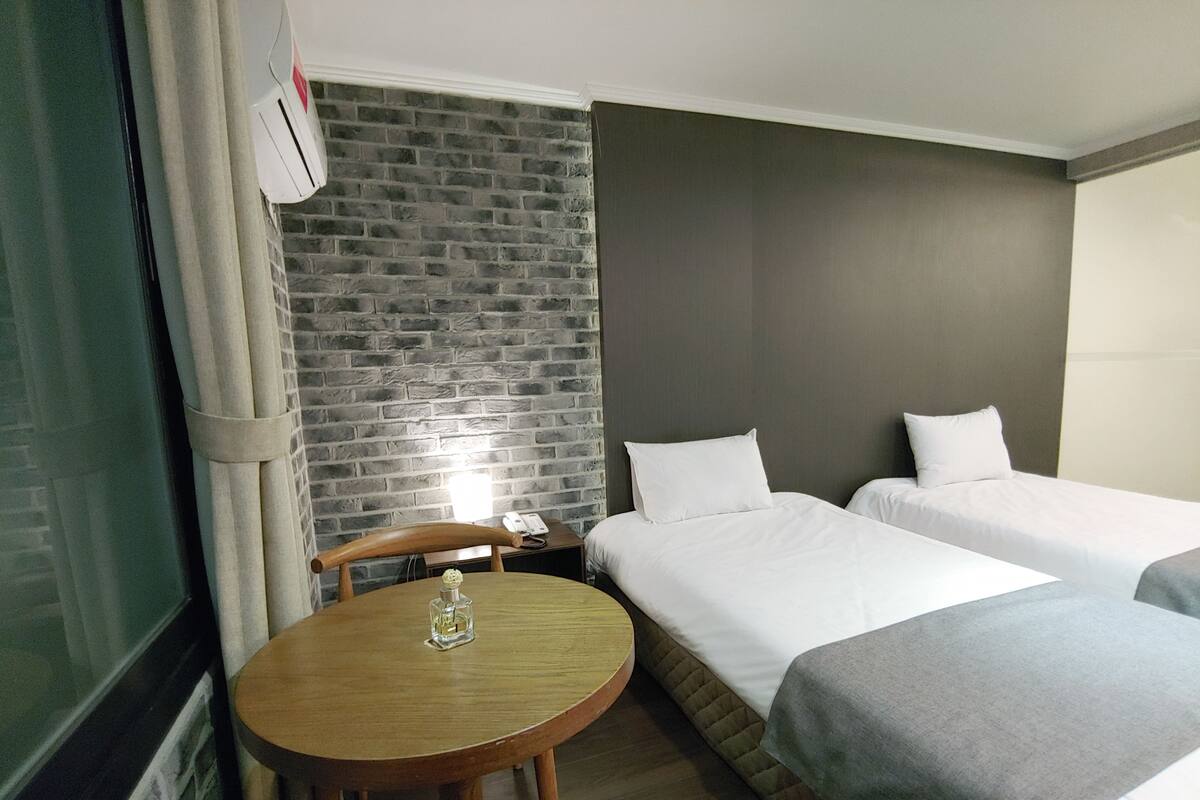 A hotel room with two single beds, a small round table with a vase, a brick-patterned wall, and a plain grey headboard.
