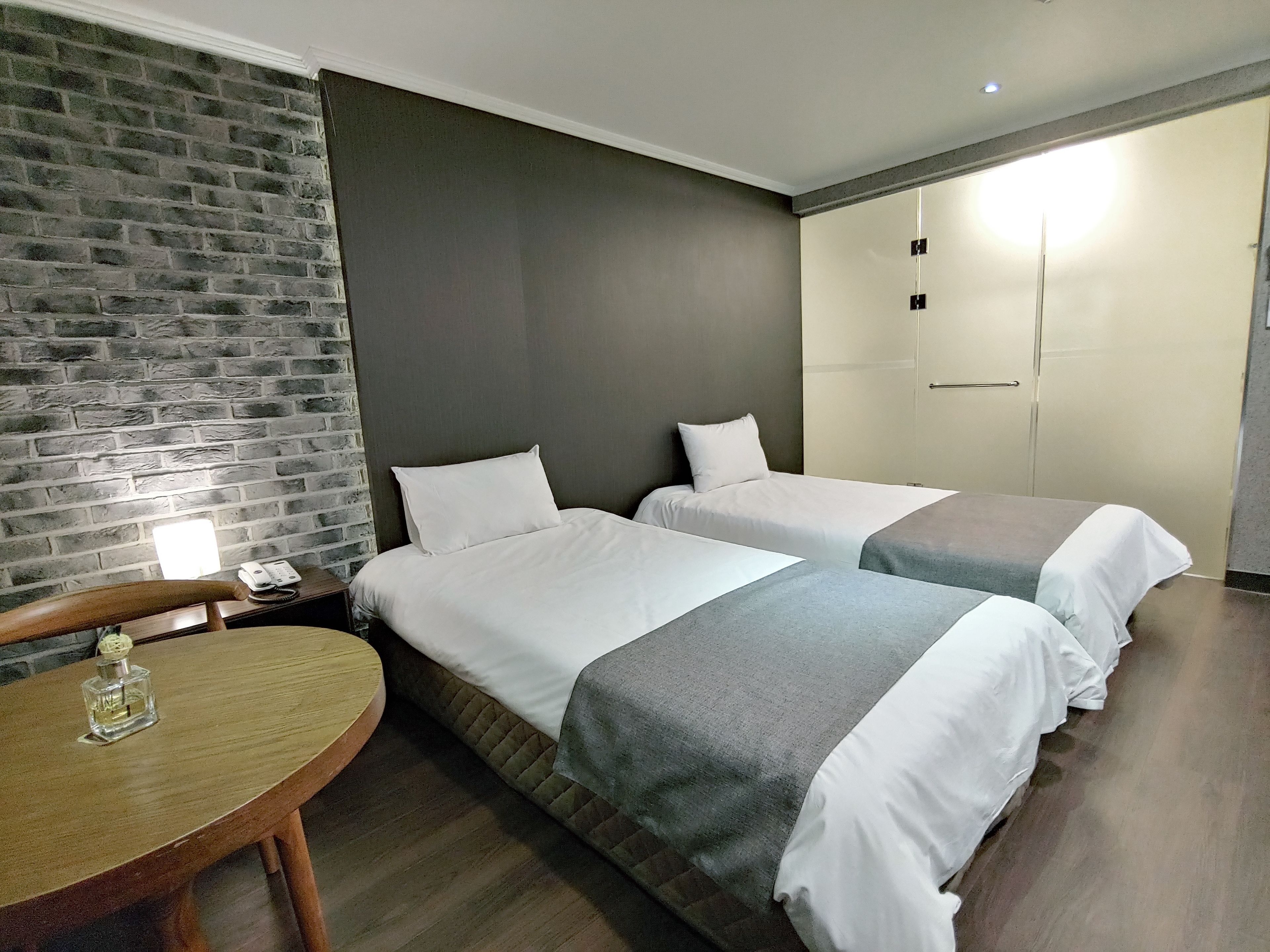 A hotel room with two beds, a wooden table, a brick wall, and a wardrobe.