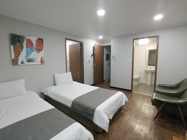 A hotel room with two beds, a painting on the wall, a green chair, and a bathroom visible through an open door.