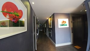 Un couloir d’hôtel moderne, avec des œuvres d’art encadrées accrochées aux murs, un ascenseur bien visible et un environnement propre et bien éclairé.