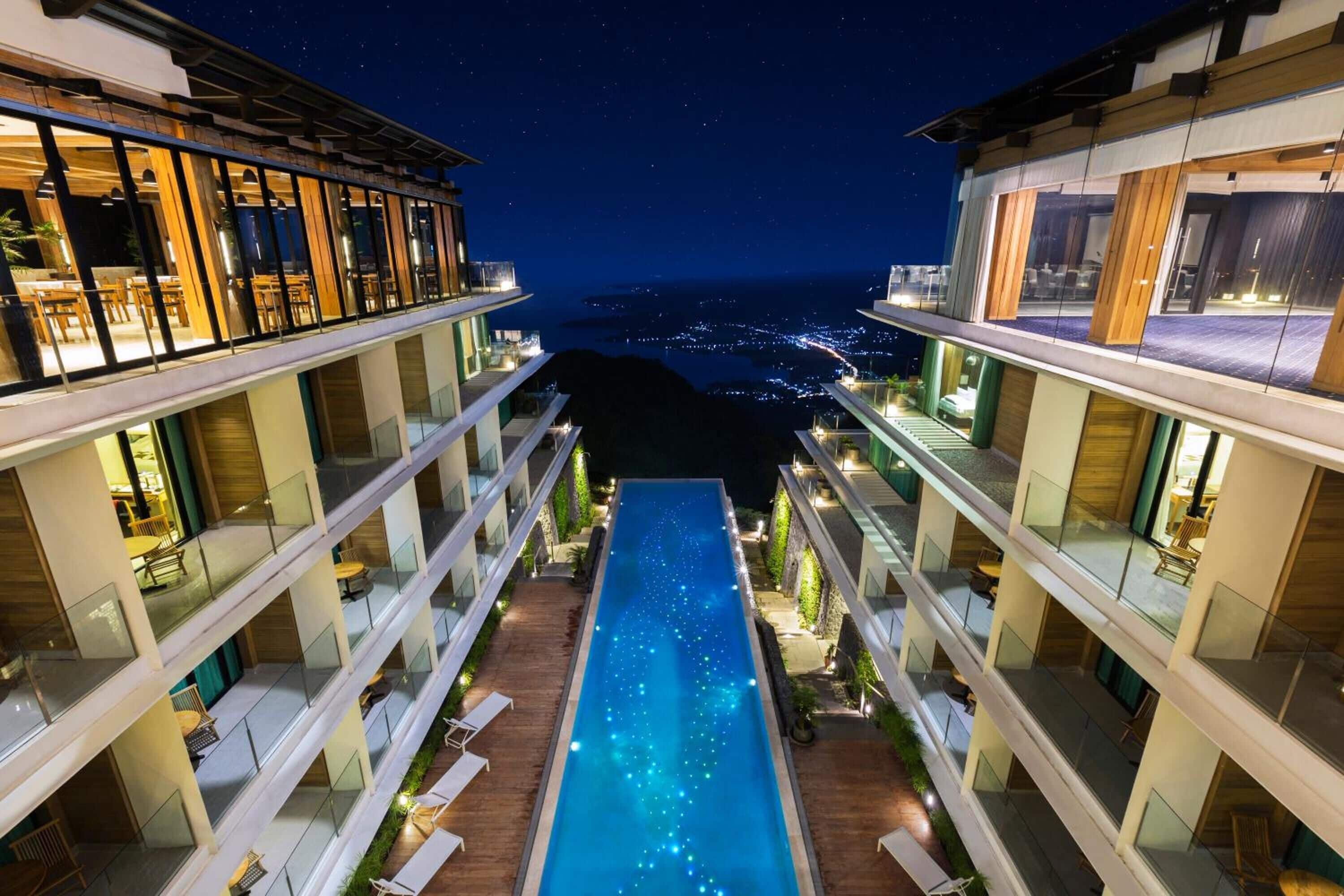 Un hôtel moderne doté d’une piscine centrale, de nombreux balcons et offrant une vue sur la ville la nuit.