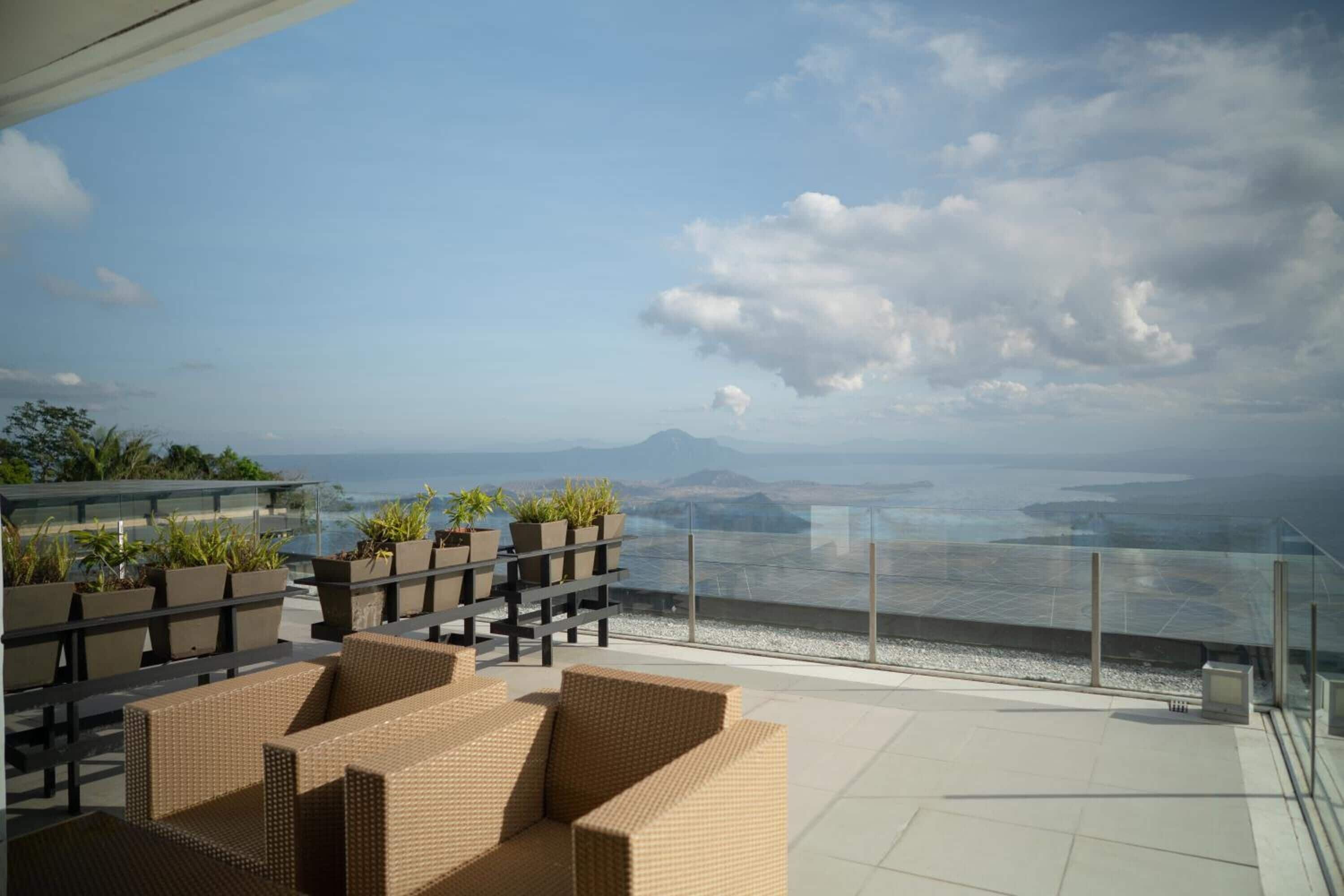 A rooftop terrace with wicker furniture, potted plants, and a glass railing overlooking a coastal view.