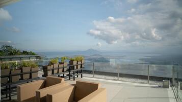 A rooftop terrace with wicker furniture, potted plants, and a glass railing overlooking a coastal view.