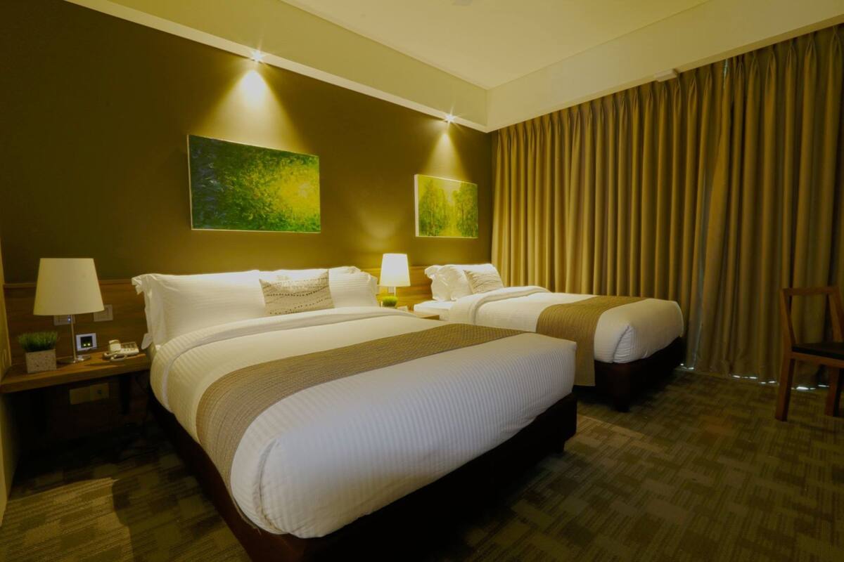 Habitación de hotel con dos camas, una mesita de noche con lámpara, una silla y un ventanal con cortinas.