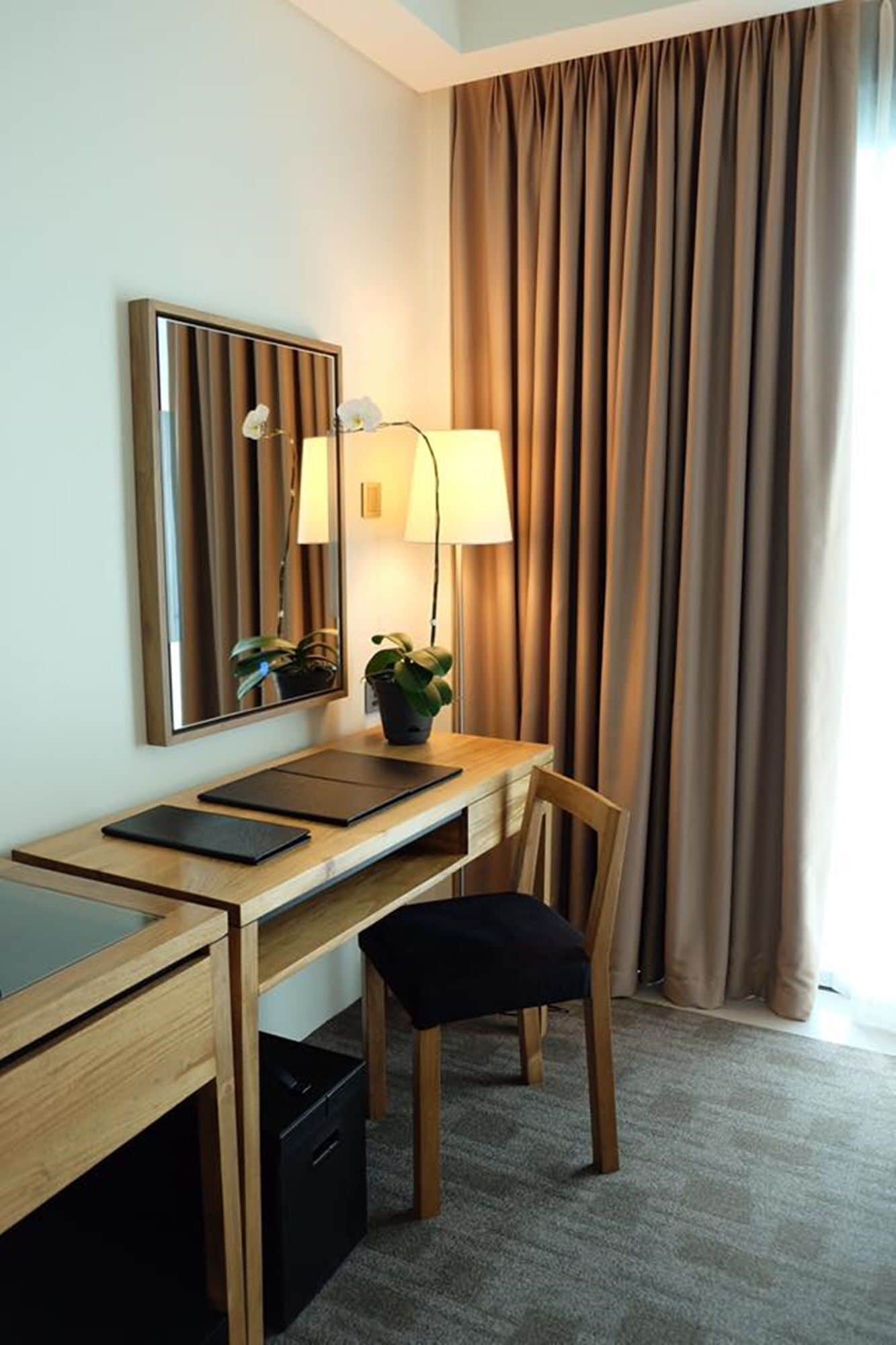 A hotel room with a wooden desk, a chair, a lamp, a mirror, and a potted plant.