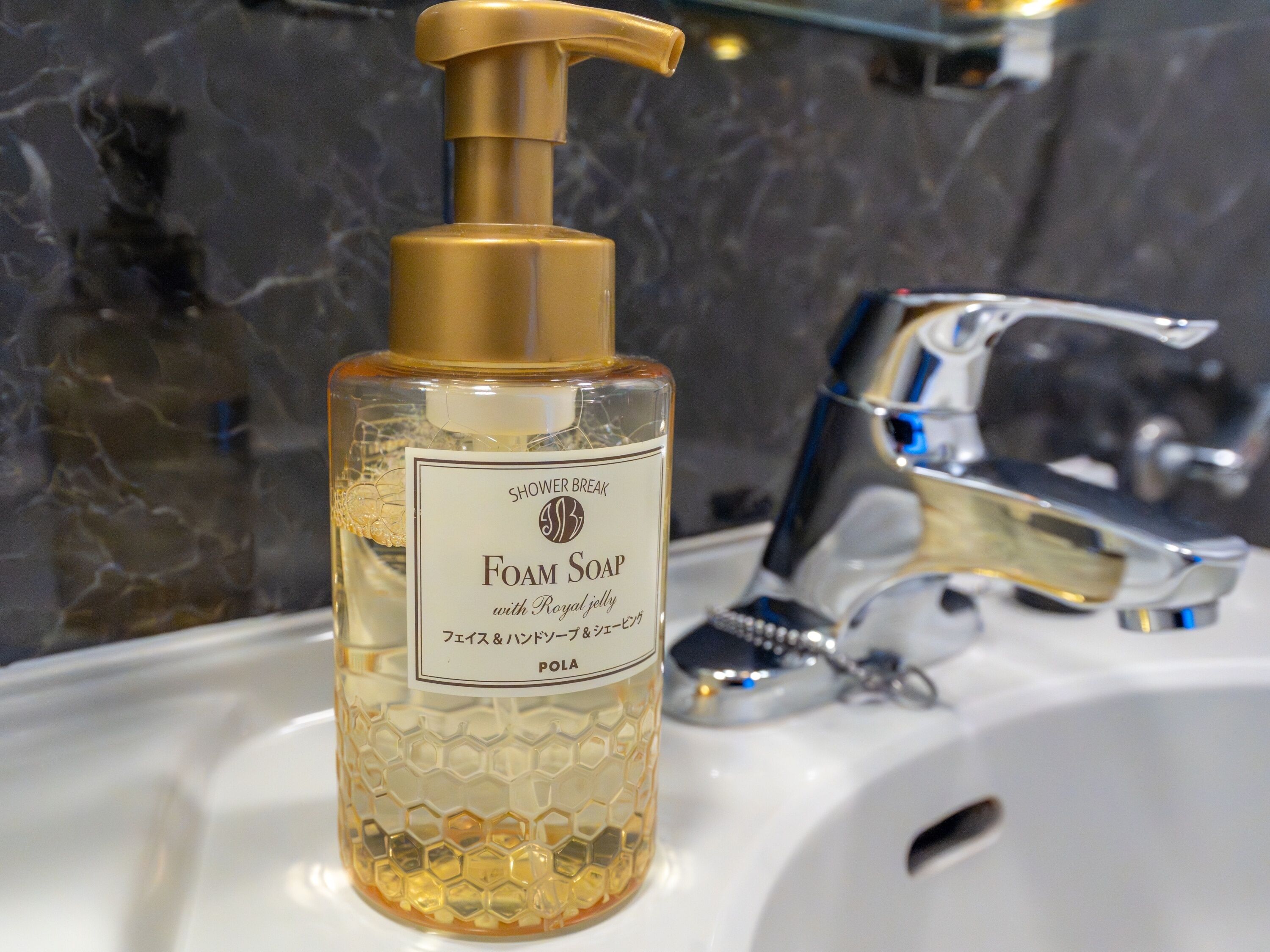 A bathroom with a clear glass bottle of foam soap with a pump dispenser, a chrome faucet, and a marble wall.
