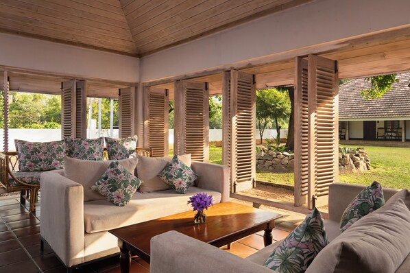 Un espace extérieur aménagé, abrité et doté d’un canapé et de fauteuils, de volets en bois et d’une vue sur une maison.
