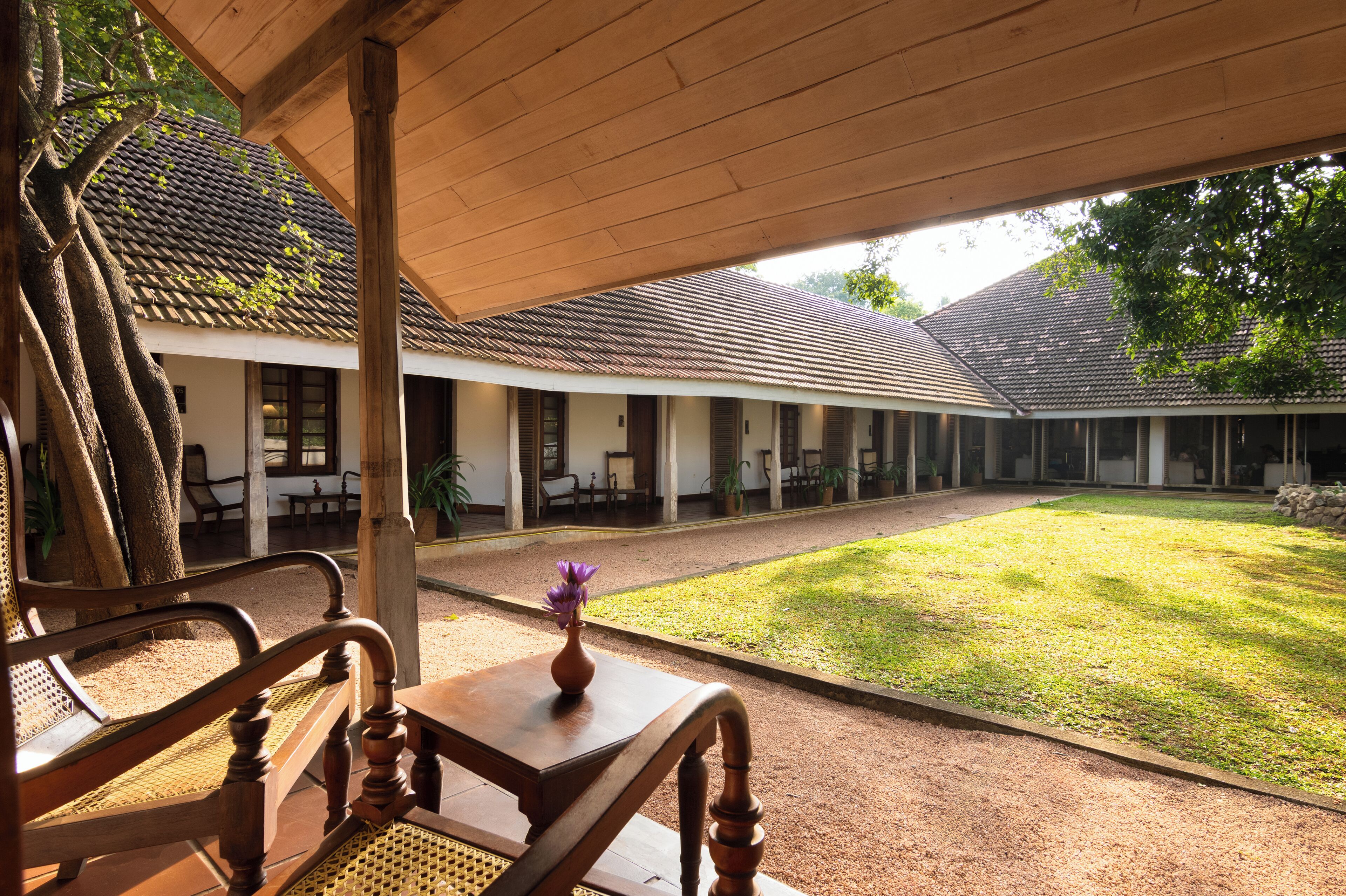 A covered outdoor area with a wooden bench and a table, a grassy courtyard, and a building with a tiled roof.