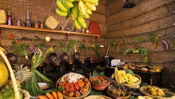 Una habitación con techo tejido, estantes con diversos objetos, y una mesa con comida y frutas.