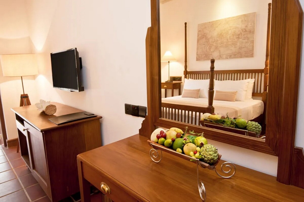 Habitación de hotel con escritorio de madera, televisor de pantalla plana, mesita de noche con fruta, cama con cabecera de madera y un mapa en la pared.