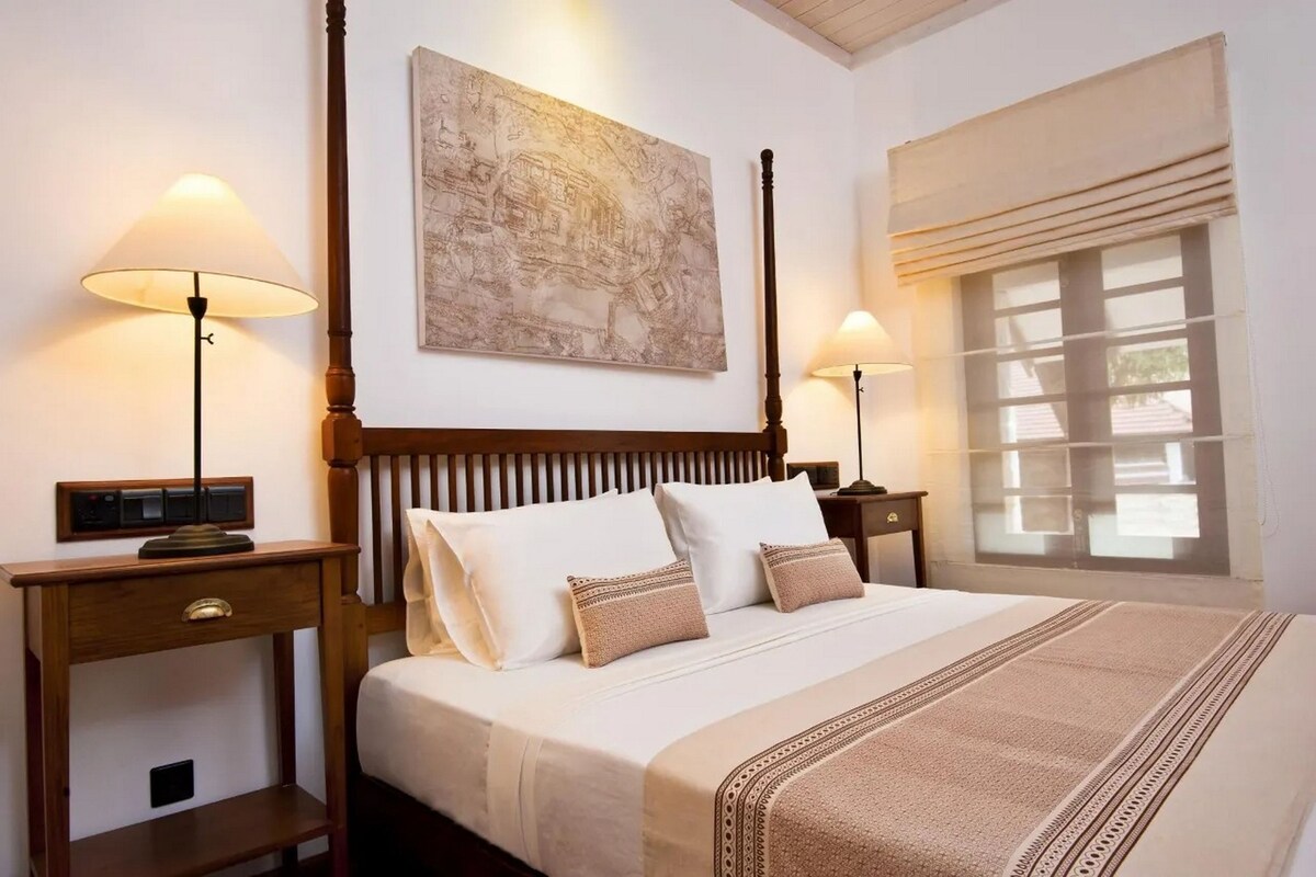 Una cama bien hecha con sábanas blancas y dos almohadas, flanqueada por mesitas de noche de madera con lámparas. Encima de la cama, se observa un mapa enmarcado de gran tamaño, y a la derecha se ve una ventana con persianas.