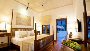 Habitación de hotel con cama con dosel, ventilador de techo, un cuadro grande, ventana con vistas y una cesta de frutas en la mesita de noche.
