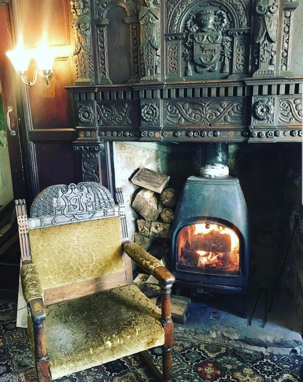 A traditional fireplace with intricate carvings, a wooden chair with a cushion, and a stone wall.