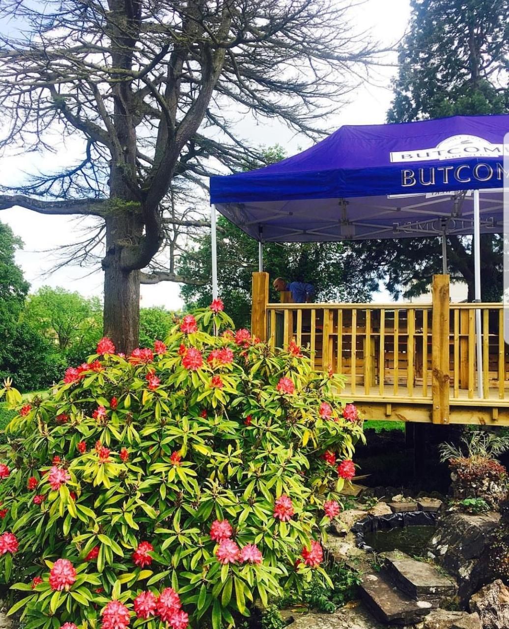 A gazebo with a wooden deck, surrounded by lush greenery and a small pond.