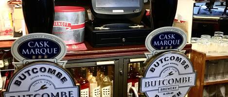 Eine Barbereich mit zwei Zapfhähnen, beschriftet mit 'Cask Marque' und 'Butcombe Bitter'. Es gibt Regale mit verschiedenen Flaschen und im Hintergrund einen Fernseher.