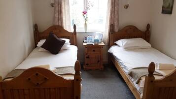 Een kamer met twee houten bedden, een klein nachtkastje met een bloevas en een raam met gordijnen.