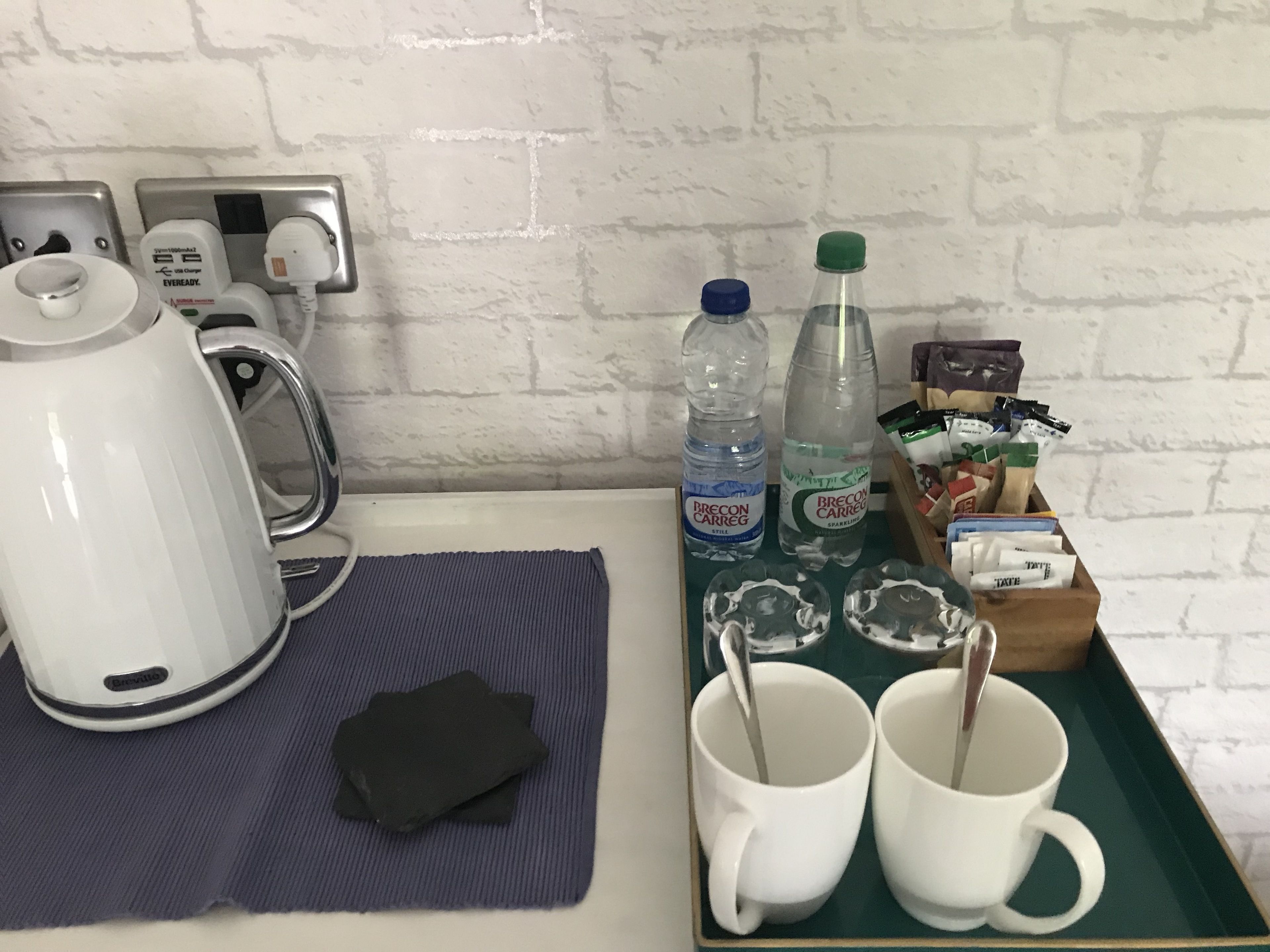 Una kitchenette con un hervidor, dos botellas de agua, una cafetera, una bandeja con dos tazas y una cuchara, y una pequeña caja de bolsitas de té.