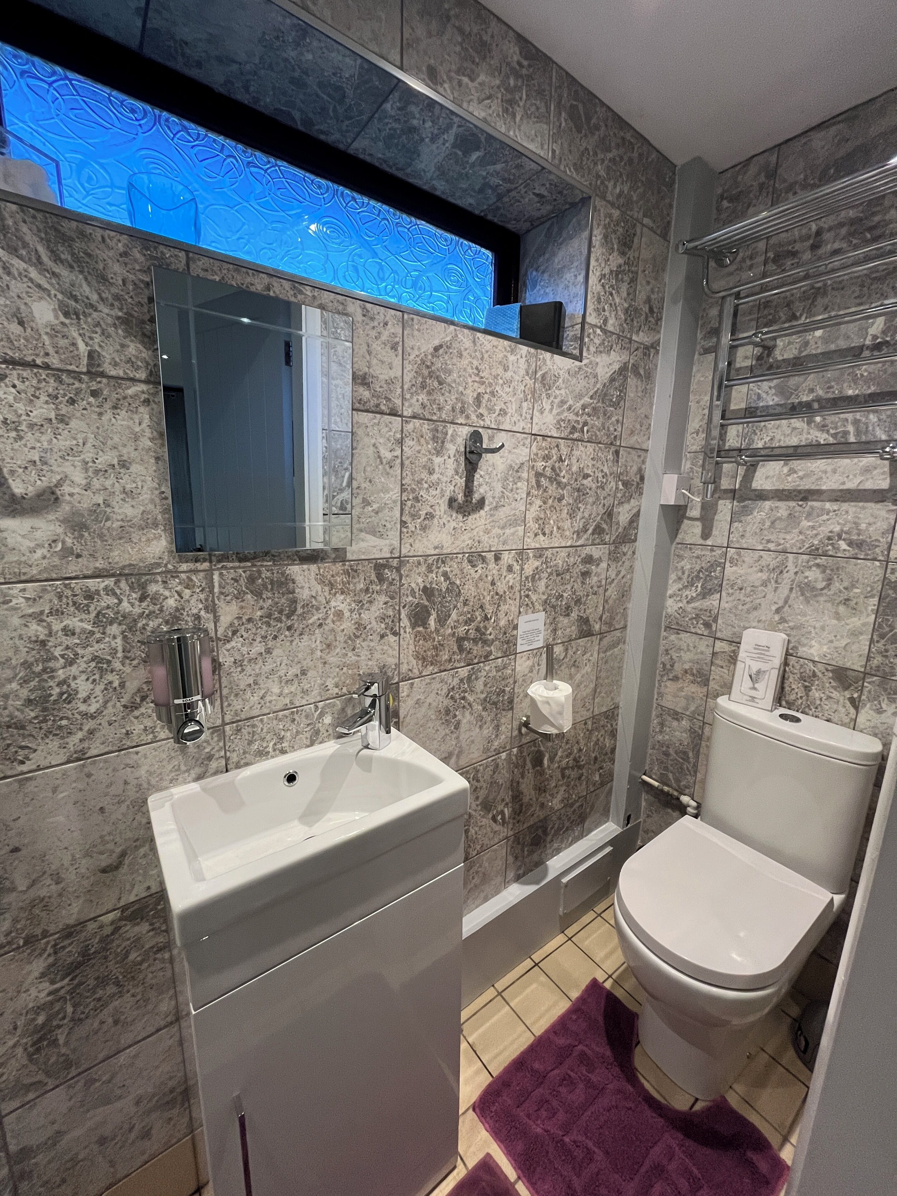 Un baño con pared de mármol, un lavamanos blanco, un inodoro y una alfombrilla de baño morada.