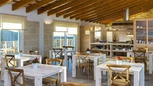 A dining area with wooden tables and chairs, a kitchen in the background, and large windows offering a view. - Agroturisme Son Vives - Adults Only (Ferreries)