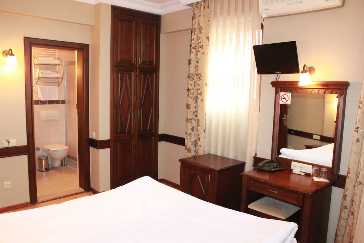 A hotel room with a bed, a wooden desk with a telephone and lamp, a television mounted above a mirror, and a bathroom visible through an open door.
