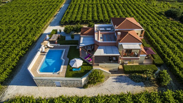 Une grande maison avec une piscine, plusieurs espaces de détente extérieurs et un vignoble en arrière-plan.