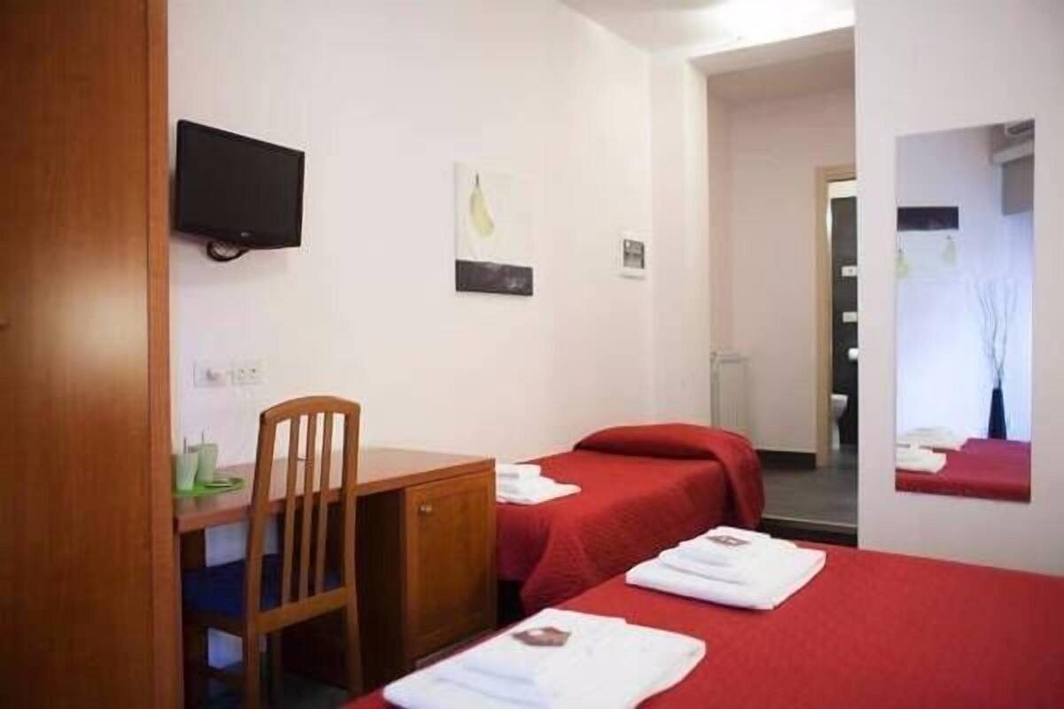 Habitación de hotel con dos camas individuales, un escritorio con silla, un televisor montado en la pared y un cuadro pequeño.