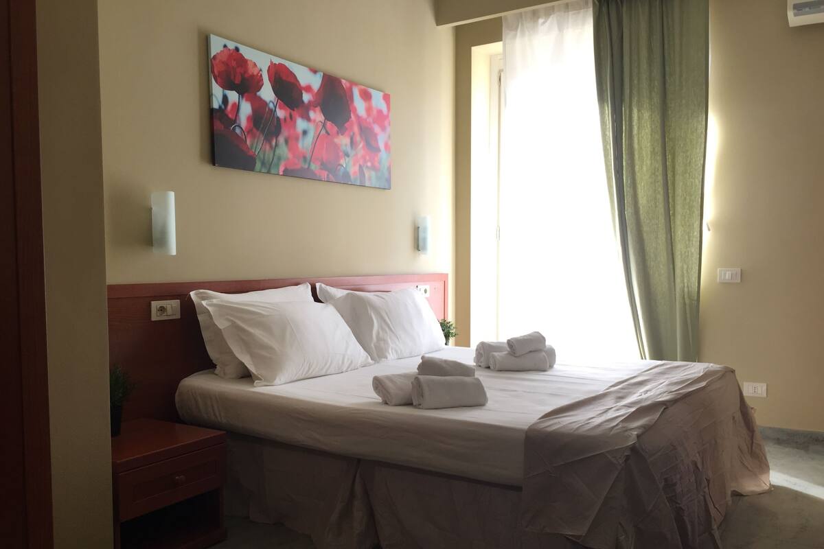 Una cama bien hecha con sábanas blancas y falda de cama color beige, una mesita de noche con una lámpara, y un cuadro de flores rojas en la pared.