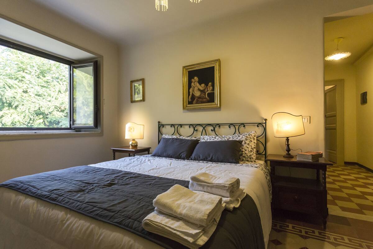 A neatly made bed with a quilt, pillows, and folded towels. There are two bedside lamps and a framed picture on the wall.