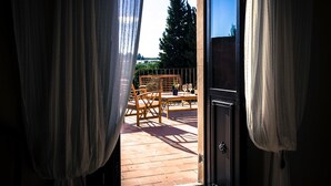 Une porte ouverte donnant sur un balcon aménagé avec du mobilier en bois, d’où l’on peut admirer un arbre et le ciel.