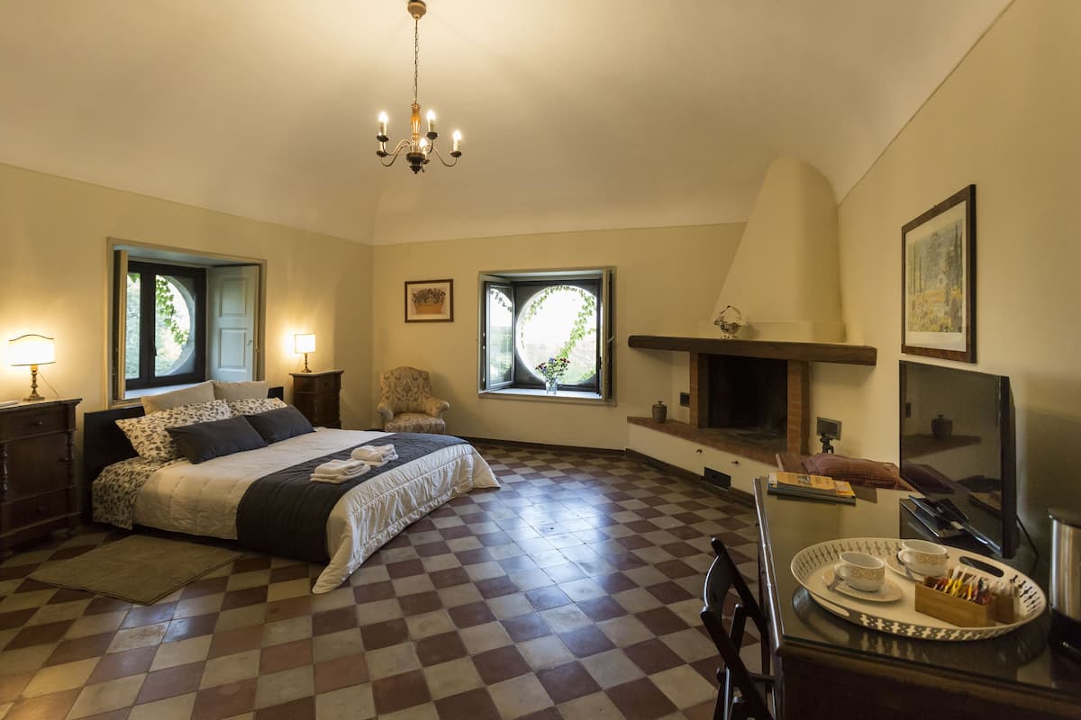 A bedroom with a checkered floor, a bed with a white and black bedspread, a chandelier, two windows with curtains, a fireplace, and a television.