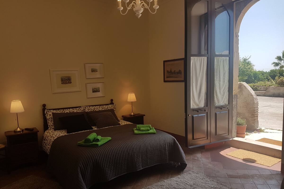 A bedroom with a bed, two bedside tables, a chandelier, and a door leading to an outdoor area.