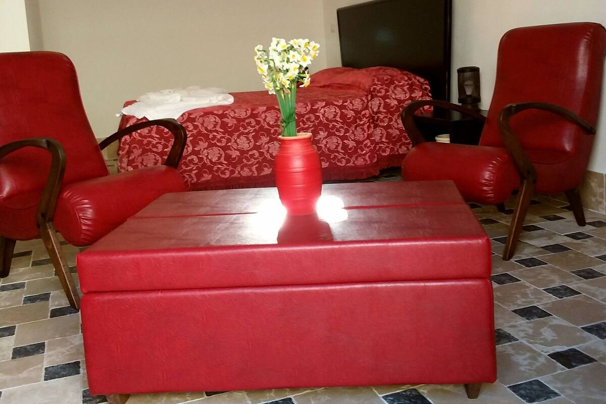 Un sofá rojo con un otoman a juego, un jarrón con flores y una cama con colcha estampada roja.