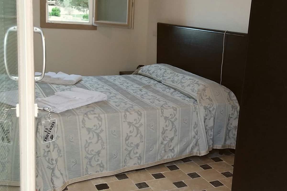 Una cama pequeña y bien hecha con una colcha estampada y almohadas, una ventana con vistas, y un suelo de baldosas.