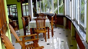 Zona de dinar amb cadires de fusta i una taula, finestres grans i un terra amb rajoles.