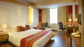Quarto de hotel com uma cama grande, uma escrivaninha com cadeira, televisão e janela com cortinas.