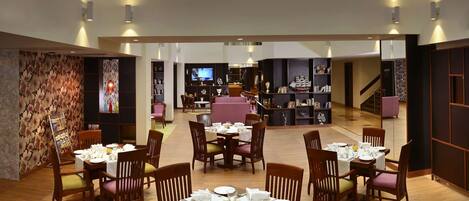 A spacious dining area with multiple tables and chairs, a central seating area with a pink sofa, and a large bookshelf in the background.