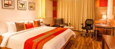 Habitación de hotel con una cama grande, un escritorio, una silla y una mesita. Hay cortinas, una lámpara y cuadros en las paredes.