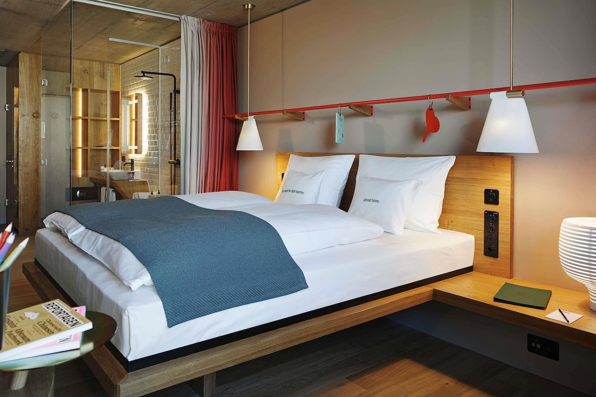 A modern hotel room with a large bed, bedside tables, a mirror, and a red accent wall.