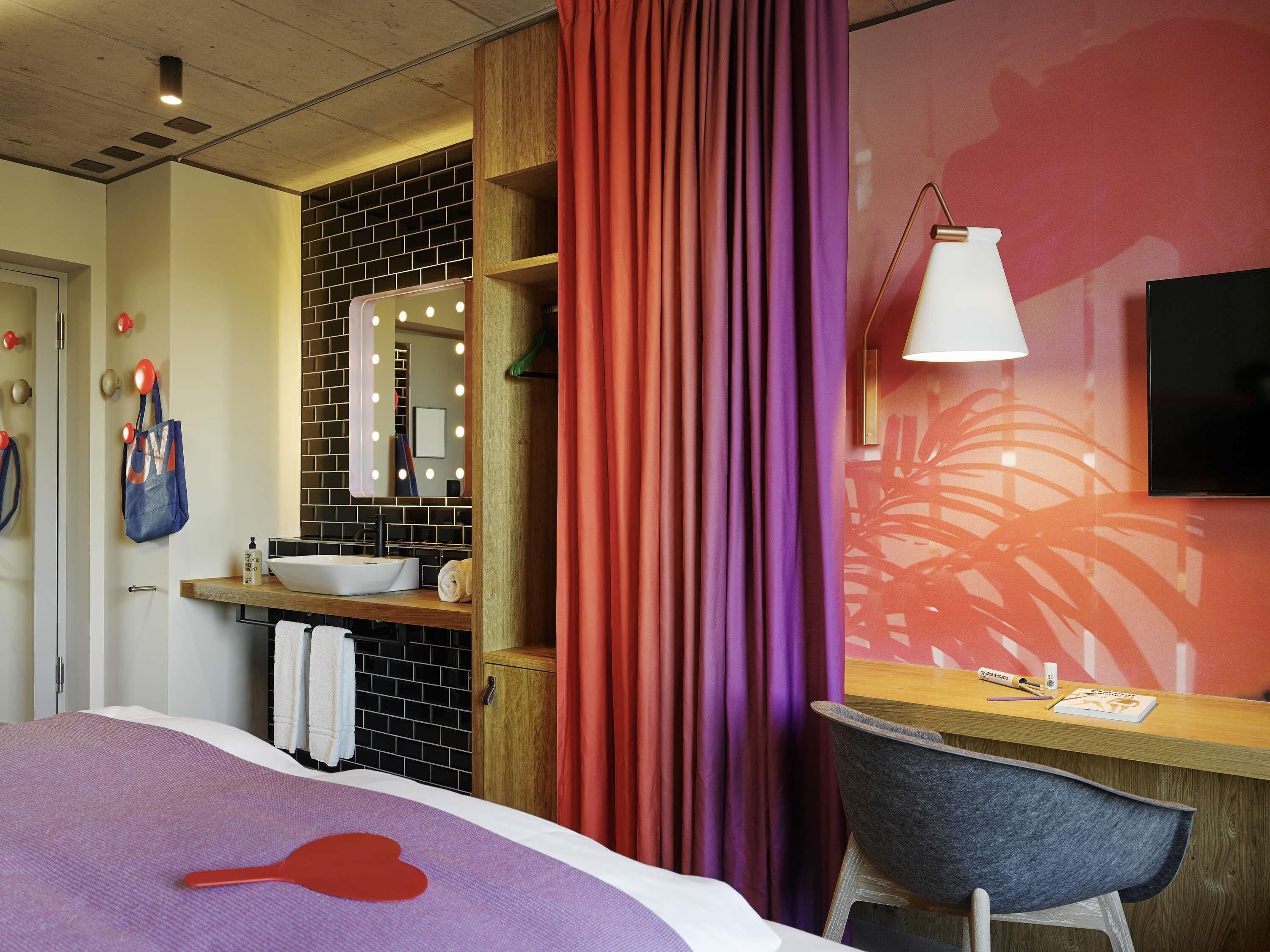 A hotel room with a bed, a desk with a chair, a bathroom with a mirror and lights, and a TV on the wall.