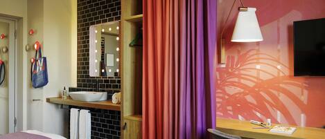 A hotel room with a bed, a desk with a chair, a bathroom with a mirror and lights, and a TV on the wall.