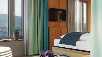 Ein modernes Hotelzimmer mit großem Fenster, einem Flachbildfernseher, einer Couch und einem kleinen Tisch mit Pflanze und Tasche.