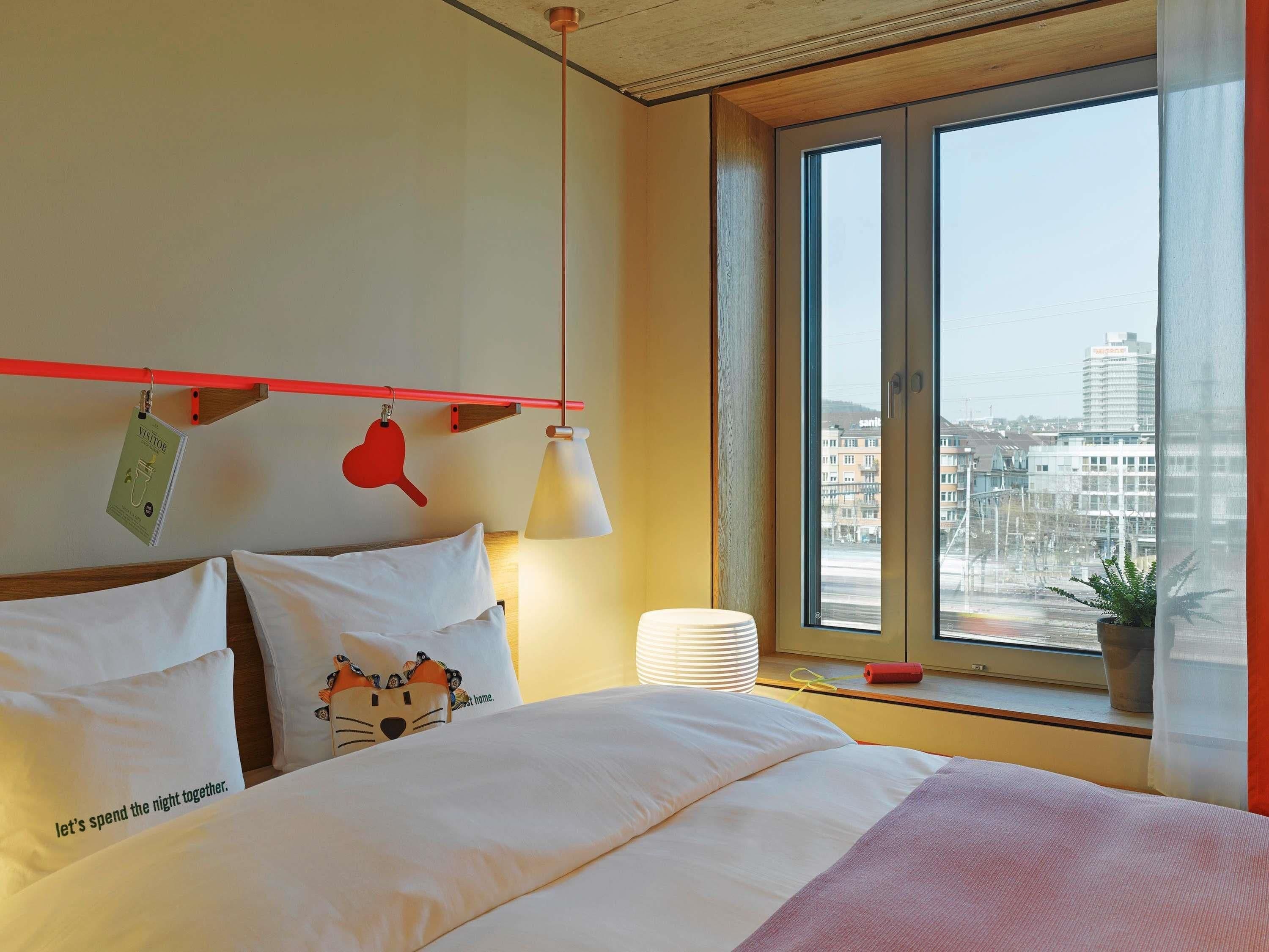 A hotel room with a bed, pillows, a hanging sign, a window with a view of buildings, and a plant on the windowsill.